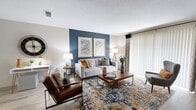 This single-floor residence in Jacksonville, Florida offers a cohesive, contemporary interior designed with both comfort and functionality in mind. Throughout the space, wood-look plank flooring provides a warm, unified foundation that flows seamlessly from room to room.

The living area serves as the focal point of the home, anchored by a bold blue accent wall that adds depth and character. Large sliding glass doors with vertical blinds and decorative curtain panels flood the room with natural light while providing access to the outdoors. A dedicated media wall accommodates a wall-mounted television above a stylish credenza.

The dining area is positioned adjacent to the kitchen, featuring a ceiling fan and a pass-through window that connects to the kitchen, enhancing the open feel between spaces. The kitchen is well-equipped with white shaker-style cabinetry, granite-look countertops, stainless steel appliances, and a window above the sink that brings in additional natural light.

The bedroom is a calm retreat with neutral tones, wood-look flooring, and a bright window dressed with navy curtains. A hallway connects the private areas of the home, leading to bathrooms that feature granite-topped vanities with white cabinetry and clean, bright finishes throughout.