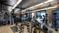 This fitness center in Dallas offers a thoroughly equipped workout environment designed to support a wide range of training goals. The space features a sleek, modern aesthetic with a dark ceiling accented by exposed ductwork, recessed lighting, and linear pendant fixtures that create a focused, energetic atmosphere. Light wood-tone flooring contrasts the darker upper elements, contributing to a clean and contemporary look.

The free weights area is prominently featured, with an extensive rack of dumbbells and multiple barbell stations loaded with weight plates, catering to strength training enthusiasts of all levels. Full-length mirrors line one wall, enhancing the sense of space and allowing users to monitor their form during exercise.

Along the perimeter, large windows allow natural light to filter in while offering views to the outdoors, creating a more open and inviting environment. The cardio section is well-appointed with a row of treadmills and other cardio machines, each equipped with individual monitors. A ceiling fan aids in air circulation throughout the space. Overall, the fitness center balances functionality with a polished, upscale design.