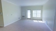 This single-floor space features a spacious, open living area that is both bright and inviting. The room is finished with soft, neutral-toned carpeting that extends throughout, providing a clean and comfortable foundation. Crown molding lines the ceiling, adding a refined architectural detail that elevates the overall aesthetic. The walls are painted in a light, neutral hue, contributing to the airy atmosphere.

Natural light floods the space through a large multi-pane window and a glass-paneled exterior door, which also offers views of an outdoor area, likely a balcony or deck. The combination of these openings ensures the room remains well-lit throughout the day. Floor-level HVAC vents are positioned along the baseboards, indicating a built-in climate control system. A wall-mounted unit is also present, providing additional temperature regulation.

The generous layout offers ample room for a variety of furniture arrangements, making it highly versatile. The clean lines, neutral palette, and thoughtful architectural detailing create a welcoming environment that is move-in ready and adaptable to a range of interior design styles.