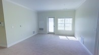 This single-floor space features a spacious, open living area that is both bright and inviting. The room is finished with soft, neutral-toned carpeting that extends throughout, providing a clean and comfortable foundation. Crown molding lines the ceiling, adding a refined architectural detail that elevates the overall aesthetic. The walls are painted in a light, neutral hue, contributing to the airy atmosphere.

Natural light floods the space through a large multi-pane window and a glass-paneled exterior door, which also offers views of an outdoor area, likely a balcony or deck. The combination of these openings ensures the room remains well-lit throughout the day. Floor-level HVAC vents are positioned along the baseboards, indicating a built-in climate control system. A wall-mounted unit is also present, providing additional temperature regulation.

The generous layout offers ample room for a variety of furniture arrangements, making it highly versatile. The clean lines, neutral palette, and thoughtful architectural detailing create a welcoming environment that is move-in ready and adaptable to a range of interior design styles.