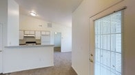 This single-floor residential unit offers a clean, functional layout designed for comfortable everyday living. The interior is finished in a bright, neutral color palette with white walls and trim that lend a fresh, airy feel throughout the space.

The kitchen is positioned along one wall and features white cabinetry with open upper shelving, white appliances including a refrigerator and range, and a peninsula counter that provides a natural division between the kitchen and the adjoining living area. The counter offers additional workspace and serves as a casual dining or prep surface.

The living area flows openly from the kitchen, finished with wall-to-wall carpet in a warm neutral tone that extends into the connected hallway and additional rooms beyond. The open sightlines from the entry through to the rear of the unit create a sense of depth and continuity throughout.

Vaulted ceilings rise noticeably above the main living and kitchen areas, adding vertical volume and contributing to the overall sense of openness. Natural light enters through a windowed door fitted with horizontal blinds near the entry, supplementing the overhead lighting.

A hallway visible from the main living area leads to additional rooms at the rear of the unit, completing a straightforward and practical floor plan suited to a range of occupants.
