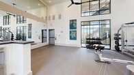 Located at 16101 Murphy Ave in Irvine, CA, this well-appointed fitness center offers a spacious, single-floor layout designed to accommodate a full range of exercise activities. The facility is immediately distinguished by its soaring double-height ceilings, which lend an open, airy atmosphere throughout the space. Floor-to-ceiling windows with sleek black frames line the walls, flooding the interior with natural light and providing views of the surrounding outdoor greenery, including palm trees characteristic of the Southern California setting. Ceiling fans are thoughtfully positioned throughout to maintain comfortable airflow.

The exercise floor is covered in durable, textured carpet tile and is organized into distinct functional zones. A robust cardio section houses multiple treadmills, elliptical trainers, stair climbers, and rowing machines, all equipped with individual digital consoles. The strength training area features a comprehensive selection of cable machines, multi-station weight systems, adjustable benches, and a well-stocked dumbbell rack. Structural columns are integrated seamlessly into the layout, preserving an open feel while defining different workout areas. Colorful abstract artwork adorns the white walls, adding a vibrant, modern aesthetic. Large wall-mounted displays and mirrors further enhance the functional and visual appeal of this well-equipped fitness facility.