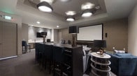 This single-floor entertainment space is designed with a sleek, contemporary aesthetic that blends function and style seamlessly. The centerpiece of the room is a large projection screen mounted on a textured neutral-toned wall, creating an immersive viewing experience ideal for gatherings and screenings. A dark, bar-height communal table runs through the heart of the space, flanked by upholstered high-back chairs that invite guests to settle in comfortably.

The ceiling is a standout architectural feature, showcasing a series of large, drum-pendant light fixtures arranged across a coffered layout. Their warm, diffused glow creates an ambient atmosphere that is both inviting and refined. Recessed lighting complements the pendants, ensuring the space is evenly and elegantly lit throughout.

To one side, a lounge area offers plush seating and accent tables adorned with sculptural décor, adding a sophisticated residential feel to the space. Tall, dark-toned table lamps with stacked column bases provide additional layers of mood lighting. A secondary wall-mounted display is also visible in the background, extending the room's entertainment capabilities.

The overall palette — warm grays, deep charcoals, and muted neutrals — lends the space a polished, upscale character throughout.