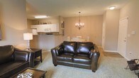 This single-floor residence offers a thoughtfully designed open-concept layout that seamlessly connects the main living areas. The living room serves as the central gathering space, featuring plush carpeting underfoot and warm, neutral-toned walls that create a welcoming ambiance. A dark leather loveseat and coordinating furniture anchor the space, complemented by an elegant chandelier in the adjoining dining area that adds a refined touch to everyday living.

The dining area accommodates a classic round table and chairs, positioned beneath the chandelier for an intimate yet functional dining experience. This space flows naturally into the kitchen, which is elevated slightly and separated by a raised breakfast bar — ideal for casual dining or entertaining. The kitchen is equipped with white cabinetry, a white appliance suite including a microwave, and ample counter space, all finished in a clean, timeless style.

Throughout the unit, the consistent use of warm beige tones and soft lighting creates a cohesive and comfortable atmosphere. The open sightlines between the living, dining, and kitchen zones make the space feel airy and connected, while the single-floor layout ensures easy accessibility and convenient day-to-day living.