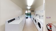 This shared laundry room is a practical, well-organized facility designed to accommodate multiple users simultaneously. The space is laid out in a long, corridor-style configuration, with rows of top-load and front-load washing machines lining both sides, complemented by a matching set of dryers. The machines are arranged along white cabinetry that provides a clean, uniform appearance throughout. Light tile flooring runs the length of the room, contributing to a bright and easy-to-maintain environment.

Overhead fluorescent lighting ensures the space is well-illuminated, and a large glass door at the far end provides both natural light and direct access to an outdoor walkway, offering a sense of openness. A ventilation unit is mounted near the exit, supporting air circulation within the room.

At one end of the facility, a small, comfortable seating area features upholstered chairs arranged around a table, accompanied by a lamp and a community bulletin board displaying informational notices. This area offers residents a convenient place to wait while their laundry cycles run. The overall ambiance is clean, functional, and welcoming, reflecting a thoughtfully maintained communal amenity.