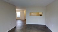 This single-floor residence offers a clean, open layout designed to maximize functionality and flow. The main living area is finished with rich, dark hardwood-style flooring that extends seamlessly throughout, providing a warm and grounded aesthetic. Crisp white walls and neutral tones create a bright, welcoming atmosphere that is both versatile and easy to personalize.

A distinctive architectural detail is the pass-through window cutout connecting the kitchen to the living space, offering convenient sight lines and an open, connected feel between the two areas. The kitchen, visible through the pass-through, features light-colored cabinetry and appears well-appointed with ample counter space and storage.

The living area opens into an adjacent room bathed in natural light from a large window, enhancing the sense of spaciousness and airiness throughout the home. The layout encourages easy movement between spaces, making the residence feel open and cohesive.

Finishing details such as clean baseboards, textured ceilings, and neutral paint tones contribute to a polished, move-in-ready presentation. Overall, this single-story residence combines practical design with comfortable living spaces, making it a well-structured home suited for a variety of lifestyles.