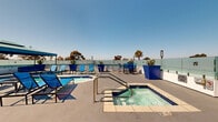 Located at 3324 Castle Heights Ave in Los Angeles, this rooftop amenity deck offers a well-appointed outdoor retreat designed for relaxation and entertaining under the Southern California sun. The space features a tiled swimming pool alongside a separate spa, both finished in classic blue tile that complements the open sky above. Generously spaced blue lounge chairs and chaise lounges are arranged throughout the sundeck, providing ample space for sunbathing and leisure. A shaded cabana structure with a teal canopy anchors one section of the deck, offering a cool retreat from direct sun, while a built-in bar station with a bold blue base provides a convenient serving area for gatherings. Frosted glass perimeter fencing maintains an open, airy feel while offering wind protection and unobstructed panoramic views of the Los Angeles skyline, surrounding palm trees, and distant mountain ranges. Elevated planters filled with succulents and tropical foliage add vibrant greenery throughout the space. Bar-height seating and bistro-style table sets provide additional areas for socializing. The overall design aesthetic is modern and cohesive, with a consistent blue-and-white color palette that evokes a resort-like atmosphere reflective of the best of Los Angeles outdoor living.