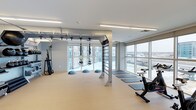 This contemporary fitness studio is designed to support a wide range of exercise modalities in a bright, open environment. The space is anchored by a centrally mounted multi-functional rig system, which supports TRX suspension trainers and provides attachment points for various functional training exercises. BOSU balance trainers are arranged along the floor, alongside a row of exercise mats ideal for yoga, stretching, or floor-based workouts. A stationary bike positioned near the windows rounds out the cardio offerings, while additional equipment such as a cable or resistance machine is visible along one wall. The studio is flooded with natural light through expansive floor-to-ceiling windows that line two walls, offering open views of the surrounding urban environment. The clean, neutral color palette — featuring light gray walls, a warm beige floor, and white ceilings with recessed linear lighting — creates an airy and motivating atmosphere. The flush, modern lighting ensures the space is equally functional during evening hours. The overall design balances functionality with a minimalist aesthetic, making it a versatile and inviting environment for individual or group fitness activities.