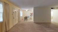 This single-floor unit offers a practical and comfortable layout with a clean, neutral aesthetic throughout. The living area is generously sized, featuring wall-to-wall carpet in a warm beige tone, cream-colored walls, and a textured ceiling. A front entry door with glass panel inserts welcomes natural light into the space, complemented by a window dressed with sheer curtains. A ceiling light fixture provides overhead illumination, and the open layout allows for flexible furniture arrangements.

The living space flows toward a separate dining area adjacent to the kitchen, where tile flooring transitions from the carpeted zones. The kitchen is equipped with white cabinetry, a coil electric range, an overhead vent hood, and light granite-style countertops that extend to a peninsula, offering additional prep and serving space. A window in the dining area brings in natural light, enhancing the bright, airy feel.

The bedroom is a well-proportioned room with plush carpeting, a ceiling fan with light fixture, and a window with curtains providing natural light. Two closets with bi-fold doors offer ample storage. The overall ambiance of the unit is light, neutral, and move-in ready.