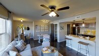 This well-appointed single-floor residence offers a thoughtfully designed open-concept layout that effortlessly connects the living, dining, and kitchen areas into one cohesive and functional space. Rich dark hardwood floors run throughout, providing a striking contrast against the neutral gray walls and crisp white trim. The living area is anchored by a comfortable seating arrangement centered around a reflective coffee table, with a ceiling fan overhead adding both style and practicality. Adjacent to the living space, the dining area features an elegant table with upholstered seating and is accented by a set of contemporary art pieces mounted on the wall, along with track lighting that highlights the décor. A built-in closet or storage space is conveniently tucked between the living and kitchen zones. The kitchen is defined by a breakfast bar with stone countertops and barstool seating, pendant lighting, and stainless steel appliances, including a full-sized refrigerator. Dark cabinetry adds depth and sophistication to the kitchen's design. The overall ambiance of the space is modern and cohesive, blending warm tones, contemporary fixtures, and an open flow that makes the area feel welcoming and versatile.