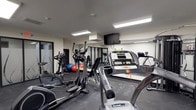 This fitness center offers a well-organized workout space designed to accommodate a range of exercise routines. The room features dark rubberized flooring, which provides both durability and traction during workouts. Overhead fluorescent panel lighting ensures the space is brightly and evenly lit throughout.

The cardio section is well-equipped with SportsArt-brand treadmills and elliptical trainers, positioned to allow comfortable use. A wall-mounted flat-screen television is installed at an elevated position, visible from the cardio equipment, providing entertainment during exercise sessions.

A mirrored wall along the back of the room enhances the sense of space and allows users to monitor their form. In front of the mirrors, a free weight and strength training area is visible, offering dumbbells and additional equipment for resistance training. Stability balls add versatility for core and flexibility exercises.

The neutral color palette—featuring white walls and grey tones—contributes to a clean, functional atmosphere. The space is efficiently arranged to maximize usability without feeling cluttered, making it a practical facility for daily fitness needs.