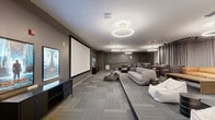 Located at 444 W College Ave in Tallahassee, Florida, this dedicated screening lounge offers a generously sized, single-floor entertainment space designed for a premium cinematic experience. The room is anchored by a large projection screen mounted on a deep gray accent wall, complemented by a media console displaying movie posters that reinforce the theatrical atmosphere. The space is finished with patterned carpet tiles throughout, contributing to both the acoustic quality and the modern aesthetic.

Seating is arranged across two levels: a main floor area and a raised platform toward the rear, providing tiered viewing angles. The main floor features a tufted gray sofa, bean bag chairs, and a leather ottoman, while the elevated section accommodates plush armchairs, a sectional sofa in warm tan leather, and additional side tables. Dramatic floor-to-ceiling drapes line the back wall, enhancing the room's immersive, cinema-like feel.

Overhead, a series of circular halo pendant lights are evenly distributed across the ceiling, casting a warm, ambient glow throughout the room. A ceiling-mounted projector is visible toward the rear of the space. The overall design palette of warm grays, taupe walls, and mixed-texture furnishings creates a sophisticated yet inviting environment well-suited for group screenings and social gatherings.
