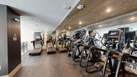 This fitness center is a thoughtfully designed workout space that maximizes its footprint with a smart, efficient layout. The room is divided into distinct cardio zones, with a row of treadmills equipped with individual monitor screens positioned along one side, and a lineup of elliptical/stair-climbing machines along the other. The slatted wood ceiling panels add a warm, contemporary aesthetic, complementing the recessed lighting that keeps the space well-illuminated throughout. Full-length mirrors line the walls, visually expanding the room and allowing users to monitor their form during exercise. The flooring transitions between rubber and carpet in different zones, providing appropriate surfaces for various workout activities. A rowing machine and stationary bike are positioned in a secondary area near a decorative iron railing, which borders an open stairwell. Large windows in this section introduce natural light and views of the surrounding greenery, enhancing the overall atmosphere. The walls feature a neutral gray and white color palette, maintaining a clean, modern aesthetic. The space is accessed via a staircase with stylish geometric iron railings, contributing to the facility's polished, upscale character.