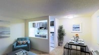 This single-floor residence offers a thoughtfully designed open-concept layout that seamlessly connects the living, dining, and kitchen areas. The kitchen is framed by a distinctive pass-through window cutout in the wall, providing visual connectivity to the main living space while maintaining a defined cooking zone. It features dark-toned cabinetry, a white refrigerator, a white range with oven, and a single-basin sink beneath the pass-through, all set against light-colored walls that keep the space feeling bright and clean. The dining area sits adjacent to the kitchen, accommodating a dining table and chairs, and is accented by wall art and a decorative potted plant that adds a touch of greenery. The living area flows naturally from the dining space, featuring plush carpeted flooring and room for comfortable seating. Neutral wall tones and textured ceilings are consistent throughout, contributing to a cohesive and calm ambiance. Flush-mount ceiling lighting provides even illumination across each area. The overall layout is efficient and practical, making excellent use of the available footprint while maintaining an open, airy feel suitable for everyday living.