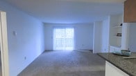 This single-floor space features an open living area characterized by clean white walls and soft, neutral-toned carpeting that extends throughout the room. The most prominent feature is a sliding glass door dressed with vertical blinds, which serves as the primary source of natural light and offers a view of the outdoors. The blinds allow for adjustable light control while maintaining privacy. The room's layout is notably open, creating a spacious and airy feel. A partial countertop or bar surface is visible along the right edge of the space, suggesting proximity to a kitchen or dining area and hinting at a functional, connected floor plan. The ceiling is smooth and uniform, complementing the minimalist aesthetic of the white walls. The overall ambiance is clean and neutral, providing a blank canvas that accommodates a variety of furniture arrangements and interior styles. The natural light from the sliding glass door enhances the brightness of the room, making it feel welcoming and open.