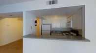 This single-floor space is designed with practicality and efficiency in mind. The kitchen serves as a central highlight, featuring a peninsula with a speckled granite-style countertop that opens toward the adjacent living area, creating a natural flow between spaces. White upper cabinetry provides ample storage, while dark lower cabinets add a subtle contrast. Appliances include a stainless steel refrigerator, an over-the-range microwave, and an electric coil range, offering a complete cooking setup. The kitchen's layout is compact yet well-organized, making it highly functional for daily use. A textured ceiling and overhead HVAC vent contribute to a standard residential feel, while warm yellow accent walls in the background add a touch of color to the otherwise neutral palette. The open-plan connection between the kitchen and the living area enhances the sense of space, allowing light to move freely throughout. Overall, the space presents a straightforward and practical layout suited for comfortable everyday living.