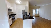 This well-appointed single-floor residence offers a thoughtfully designed open-concept layout that seamlessly connects the kitchen and living areas. The kitchen is a standout feature, equipped with stainless steel appliances — including a range, microwave, and refrigerator — and anchored by a peninsula with striking dark granite countertops and an undermount sink. Crisp white cabinetry provides ample storage, complemented by a louvered-door closet that adds a classic touch. Warm hardwood-style flooring flows throughout the space, creating a cohesive and inviting atmosphere. Crown molding adds an elegant architectural detail to the living area, which benefits from natural light streaming through a set of glass-paneled doors leading to an outdoor area. The open living space offers a versatile canvas, with generous room to arrange seating and furnishings to suit any lifestyle. The clean white walls and bright flush-mount lighting ensure the interior feels airy and welcoming. Overall, the space balances modern finishes with functional design, making it an ideal environment for both everyday living and entertaining.
