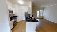 This well-appointed single-floor residence offers a thoughtfully designed open-concept layout that seamlessly connects the kitchen and living areas. The kitchen is a standout feature, equipped with stainless steel appliances — including a range, microwave, and refrigerator — and anchored by a peninsula with striking dark granite countertops and an undermount sink. Crisp white cabinetry provides ample storage, complemented by a louvered-door closet that adds a classic touch. Warm hardwood-style flooring flows throughout the space, creating a cohesive and inviting atmosphere. Crown molding adds an elegant architectural detail to the living area, which benefits from natural light streaming through a set of glass-paneled doors leading to an outdoor area. The open living space offers a versatile canvas, with generous room to arrange seating and furnishings to suit any lifestyle. The clean white walls and bright flush-mount lighting ensure the interior feels airy and welcoming. Overall, the space balances modern finishes with functional design, making it an ideal environment for both everyday living and entertaining.