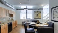 This two-floor space showcases a distinctive industrial-modern aesthetic, highlighted by exposed metallic ductwork running along the bright white ceiling of the main level. The open-concept layout integrates the living, dining, and kitchen areas into one cohesive space, promoting a sense of openness and fluid movement throughout.

The kitchen is positioned along one wall, featuring warm wood cabinetry with ample storage and dark countertops, offering a functional workspace that transitions naturally into the dining area. A round dark dining table with black leather chairs serves as a central gathering point, ideal for both casual and formal occasions.

The living area benefits from large windows that flood the space with natural light, creating an airy and welcoming atmosphere. Hardwood flooring throughout the main level adds warmth and continuity, complementing the industrial elements above.

A staircase visible from the main area leads to the second floor, suggesting additional private living spaces. The combination of exposed mechanical features, contemporary furnishings, and abundant natural light gives this multi-level space a modern, urban character.