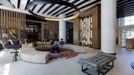 This single-floor space showcases a striking contemporary interior defined by thoughtful architectural details and high-end finishes throughout. The expansive main living area draws immediate attention with its dramatic ceiling design, featuring bold dark exposed beams arranged in a geometric pattern that contrasts sharply with the bright white surfaces below. A sculptural circular chandelier with warm amber lighting serves as a stunning focal point, anchoring the space with elegance.

The seating arrangement is centered around a generous curved sectional sofa in neutral tones, complemented by a bold rust-and-blue patterned area rug that adds a vibrant artistic element underfoot. A sleek glass coffee table and additional tufted seating in cool gray tones complete the refined furniture ensemble.

Vertical slatted wood partitions serve as architectural dividers throughout the space, creating visual depth while maintaining an open, fluid layout. An illuminated decorative screen wall, featuring backlit rectangular panels, adds warmth and a layered glow to the interior. Warm wood-toned wall cladding introduces natural texture, balancing the contemporary design elements. Overall, the space exudes a polished, modern ambiance suited to sophisticated entertaining.
