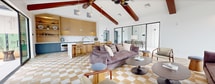 Located at 85 Livingston Ave in Roseland, NJ, this single-floor amenity space spans 1,560 square feet across three distinct areas designed for community gathering and entertainment. The centerpiece is a dramatic lounge featuring soaring vaulted ceilings with rich exposed wood beams and striking mid-century modern sputnik chandeliers. A bold gold-and-white checkered tile floor ties the space together, creating a sophisticated visual anchor throughout. Floor-to-ceiling black-framed windows flood the interior with abundant natural light while offering views of the surrounding residential community. The lounge is appointed with plush velvet seating, eclectic accent chairs, and curated greenery, lending a warm yet refined atmosphere. Adjacent to the lounge, a built-in kitchenette features light wood cabinetry, a marble-topped island with bar seating, open display shelving, and blue accent cabinetry for a designer touch. Glass partition walls connect the interior to an enclosed outdoor courtyard, which continues the checkered tile motif and offers casual wicker seating alongside a stainless steel BBQ station and communal dining table — ideal for outdoor entertaining. Together, these spaces deliver a cohesive, upscale amenity experience.
