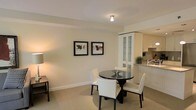 This well-appointed single-floor space presents a seamlessly integrated layout that combines a dining area and kitchen in an open-plan arrangement. The dining space is anchored by a sleek, round dark-glass table surrounded by upholstered cream chairs with nailhead trim detailing, lending a refined, classic touch to the room. A white display cabinet with glass-fronted shelving stands as a prominent feature, offering both storage and an elegant focal point.

The walls are finished in soft, warm neutral tones that create a calm and inviting atmosphere, complemented by light-colored carpeting that adds texture underfoot. Two framed botanical art pieces adorn the wall, adding subtle visual interest without overwhelming the understated aesthetic.

The adjoining kitchen is compact yet functional, featuring clean white cabinetry, light stone countertops, and modern appliances including a microwave. A single-basin sink is positioned beneath cabinetry, maximizing the use of available space. Recessed and flush-mounted ceiling lighting provides consistent illumination throughout, enhancing the bright and airy feel of the interior. Overall, the space conveys a polished, comfortable environment with a cohesive design sensibility throughout.