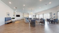 This generously proportioned single-floor community space is designed with a coastal aesthetic, blending crisp white walls, light wood-tone flooring, and a consistent navy and white color palette throughout. The open-concept main lounge accommodates multiple seating arrangements, including plush sofas, armchairs with woven rope-style bases, and intimate conversation groupings, all anchored by recessed lighting and elegant pendant chandeliers overhead. A wall-mounted flat-screen TV serves as a focal point in the lounge area, complemented by decorative starburst wall accents nearby.

The kitchenette area features white shaker-style cabinetry, a quartz countertop bar with navy upholstered barstools, a stainless steel sink, and appliances—ideal for hosting events or casual gatherings. Adjacent dining areas are bathed in natural light streaming through large windows dressed with geometric-patterned curtains and wooden blinds, offering views of lush outdoor greenery. A separate, more intimate dining nook provides a cozy round table setting with patterned upholstered chairs.

Additional seating alcoves with armchairs and small accent tables offer quieter retreats. Shiplap wainscoting, framed blue abstract artwork, and sunburst wall sconces add refined decorative touches, while glass doors lead to an outdoor area, further enhancing the bright and welcoming ambiance throughout.
