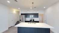 This contemporary two-story residence showcases a thoughtfully designed open-concept layout that emphasizes clean lines and modern finishes throughout. The kitchen serves as a central highlight of the main living area, anchored by a generous center island topped with a crisp white quartz-style countertop and an undermount stainless steel sink. A striking multi-pendant linear chandelier hangs above the island, providing focused task lighting that complements the recessed ceiling lights spread throughout the space. The kitchen is outfitted with a full suite of stainless steel appliances, including a French door refrigerator with water and ice dispenser, an electric range, and an over-the-range microwave. Dark, flat-panel cabinetry extends to the upper walls, offering ample storage while lending a sophisticated, modern contrast against the white backsplash and bright white walls. The flooring is finished in light-toned wood-look planks, adding warmth to the otherwise cool palette. A staircase is visible from the kitchen, connecting to the second floor of the residence. The overall ambiance is bright, clean, and contemporary, reflecting a well-appointed space designed for both functionality and modern aesthetic appeal.