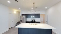This contemporary two-story residence showcases a thoughtfully designed open-concept layout that emphasizes clean lines and modern finishes throughout. The kitchen serves as a central highlight of the main living area, anchored by a generous center island topped with a crisp white quartz-style countertop and an undermount stainless steel sink. A striking multi-pendant linear chandelier hangs above the island, providing focused task lighting that complements the recessed ceiling lights spread throughout the space. The kitchen is outfitted with a full suite of stainless steel appliances, including a French door refrigerator with water and ice dispenser, an electric range, and an over-the-range microwave. Dark, flat-panel cabinetry extends to the upper walls, offering ample storage while lending a sophisticated, modern contrast against the white backsplash and bright white walls. The flooring is finished in light-toned wood-look planks, adding warmth to the otherwise cool palette. A staircase is visible from the kitchen, connecting to the second floor of the residence. The overall ambiance is bright, clean, and contemporary, reflecting a well-appointed space designed for both functionality and modern aesthetic appeal.