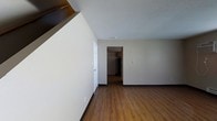 This two-story space is characterized by clean, neutral finishes and a straightforward layout designed for functional everyday living. The main level showcases warm wood-look laminate flooring that extends throughout the open living area, complemented by neutral beige walls and a textured popcorn ceiling that adds subtle dimension overhead. A staircase is visible along one wall, indicating convenient access to the upper floor. The open doorway leading toward the rear of the space suggests a connected layout, with glimpses of an additional room — likely a kitchen or utility area — visible beyond. Black baseboards provide a subtle contrast against the light-toned walls, adding a clean, defined edge to each room. The space maintains a minimalist character, offering a blank canvas that is easy to personalize. Overall, the interior presents a practical and uncluttered environment, with consistent flooring and neutral tones creating a cohesive feel throughout both the main living area and the connecting spaces visible from this vantage point.