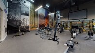 Located at 1000 Elements Way in Irvine, California, this single-floor fitness facility offers a well-rounded, thoughtfully designed environment for a wide range of workout experiences. The space is divided into distinct functional zones, each catering to different training needs.

At the heart of the facility is an expansive indoor turf field, marked with yardage lines and equipped with overhead suspension rigs, TRX systems, and functional training stations — ideal for athletic conditioning, agility drills, and group training sessions. Adjacent to the turf is a robust strength and equipment zone featuring rowing machines, cable systems, weight racks, and resistance machines, all set beneath a dramatic dark industrial ceiling with exposed ductwork and bold pendant lighting.

Striking large-scale nature murals depicting forests, canyons, and landscapes adorn the walls of the strength area, adding an immersive visual dimension to workouts. A dedicated studio space enclosed by floor-to-ceiling glass walls features warm hardwood flooring and is clearly marked for yoga and mindfulness use, with a sculptural meditation figure greeting visitors at the entrance. Signage reading "BREATHE" reinforces the calming, intentional atmosphere of this area.

Throughout the facility, recessed and linear LED lighting ensures consistent illumination, while mirrored walls enhance the sense of openness and space. The overall design balances high-performance athletic functionality with a motivating and visually engaging aesthetic.