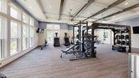 Located at 480 John Carter Rd in Savannah, Georgia, this single-floor fitness center offers a generously sized, thoughtfully designed exercise space. The room's architecture is immediately impressive, showcasing soaring vaulted ceilings with exposed rustic wood beams, coffered detailing, and recessed lighting that creates a bright, welcoming atmosphere. Large multi-pane windows line the walls, flooding the space with natural light and providing views of the surrounding greenery.

The flooring transitions between durable hardwood in the cardio and cycling area and plush carpet in the strength training zone, clearly defining separate workout areas. A full-length mirror wall expands the sense of space in the cycling section, which is equipped with stationary bikes and wall-mounted televisions. The strength training area features a robust TRX and power rack system complete with free weights, kettlebells, and medicine balls, alongside benches for versatile lifting. Cardio equipment includes stair climbers, treadmills, ellipticals, and a rowing machine. Ceiling fans throughout ensure comfortable air circulation. The space also provides access to an adjacent room, likely for changing or restrooms, making this fitness center a comprehensive and polished facility.