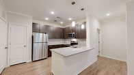 This efficient and thoughtfully designed 1-bedroom, 1-bathroom residence offers a functional single-floor layout with a cohesive modern aesthetic throughout. The open-concept kitchen is a standout feature, showcasing rich dark-brown cabinetry, granite countertops, and a full suite of stainless steel appliances including a refrigerator, range, microwave, and dishwasher. A peninsula with an integrated sink adds both workspace and a natural division between the kitchen and the living area. Light wood-look flooring runs through the main living spaces, complementing the neutral wall tones and crown molding details. The living room benefits from natural light through glass patio doors and features a ceiling fan overhead. The bedroom is carpeted for comfort, with a window allowing in natural light and access to a dedicated closet. The bathroom continues the design language of the kitchen with matching dark cabinetry, granite countertops, an undermount sink, and a large vanity mirror. A hallway connects the main living areas to the private spaces, providing a clear and practical flow throughout the residence.