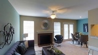 This single-floor living space centers around a well-appointed living room that combines comfort and style. The room features a wood-surround fireplace as its focal point, flanked by windows fitted with white blinds that allow natural light to filter in softly. A decorative sunburst mirror mounted above the fireplace mantel adds a striking visual element to the neutral, taupe-toned walls.

The space is finished with light carpet flooring and includes a ceiling fan with an integrated light fixture, ensuring year-round comfort. Contemporary furnishings, including a sleek black swivel accent chair and a dark sofa with decorative pillows, complement the room's modern yet relaxed aesthetic.

French doors on one side of the room introduce additional natural light and provide access to an exterior area, enhancing the sense of openness. A dining area is partially visible near the French doors, suggesting a practical, flowing layout between living and dining spaces. Overall, the room offers a balanced blend of functional design and inviting ambiance.
