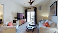 This single-floor space offers a thoughtfully designed living area that balances comfort and functionality. The living room serves as the heart of the space, featuring plush, light-toned seating arranged around a set of elegant nested round coffee tables with a dark wood and metal finish. A large flat-screen television is positioned atop a sleek, multi-toned media console, complemented by decorative greenery that adds a fresh, natural touch to the room.

Natural light flows generously into the space through a sliding glass door framed by floor-length dark curtains, which open to a private outdoor patio area — ideal for relaxing or al fresco dining. The patio is visible through the glass door and features outdoor seating, extending the livable space beyond the interior.

The living room is finished with soft wall-to-wall carpeting underfoot, contributing to a warm and inviting ambiance. Overhead, a stylish ceiling fan with a rich wood-toned finish provides both form and function, circulating air throughout the room. Neutral wall tones keep the space feeling airy and open, while warm accent lighting from a table lamp adds a cozy atmosphere. The layout is efficient and well-organized, making excellent use of the available space.