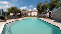 Located at 2301 Windchester St in Pearland, Texas, this single-story unit offers a thoughtfully designed interior complemented by well-maintained community outdoor amenities. The living area immediately draws attention with its soaring vaulted ceilings, which lend an open, airy quality to the space. A tile-surround fireplace with a white mantel serves as a natural focal point, adding warmth and character to the room. Dark hardwood-style flooring runs throughout, providing a rich contrast against the neutral, light-toned walls.

Large windows allow generous natural light to fill the living space, offering views of the surrounding greenery and neighborhood streetscape. The tall windows extend toward the upper portion of the vaulted ceiling in select areas, further enhancing the sense of vertical scale. A staircase with white railings and elegant arched upper windows adds architectural interest, while an in-unit laundry area is conveniently tucked beneath the stairwell.

Outdoors, the property features a well-kept grassy common area enclosed by chain-link fencing, complete with a dedicated dog park equipped with charming pet-themed metal sculptures, benches, and a fire hydrant — ideal for pet owners. The community setting is surrounded by mature trees beneath open Texas skies.