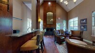 Located at 7227 Peachtree Dunwoody Rd in Sandy Springs, GA, this single-floor clubhouse at Dunwoody Ridge offers an elegant and welcoming communal environment designed with refined architectural details. The space is defined by dramatically vaulted ceilings that soar above the main gathering areas, creating an expansive, airy atmosphere throughout. Rich hardwood flooring runs continuously across the entire space, anchoring the warm, sophisticated aesthetic.

Upon entry, a stylish reception area greets visitors with a dark, nail-head-trimmed cabinet console, a decorative mirror, and ambient table lighting. French-paned glass doors frame the entrance, flooding the foyer with natural light. A bar-height counter with a green granite top provides a practical workspace or service station with barstool seating nearby.

The centerpiece of the main lounge is a floor-to-ceiling wood-paneled fireplace wall, topped by a large multi-pane mirror that reflects the chandelier above. The hearth features a dark tile surround flanked by tall decorative vases, adding a dramatic focal point to the room. Comfortable leather armchairs and accent seating are arranged in conversational groupings atop patterned area rugs, ideal for relaxed gatherings. Multiple large windows throughout invite abundant natural light and provide views of the surrounding greenery, enhancing the warm, inviting ambiance of the entire space.