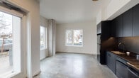 Located at 5620 N Interstate Ave in Portland, Oregon, this contemporary single-floor space showcases an industrial-modern aesthetic defined by polished concrete floors, exposed concrete columns, and crisp white walls throughout. The open living area is generously lit by multiple large windows that frame views of the surrounding neighborhood, creating a bright and airy atmosphere. A mini-split HVAC unit ensures year-round comfort, while a glass-panel front entry door with sidelights further enhances the flow of natural light into the space. The kitchen is a standout feature, outfitted with floor-to-ceiling matte black cabinetry, dark countertops, a full suite of black and stainless appliances including a refrigerator, range, and dishwasher, and under-cabinet lighting that adds a refined, modern touch. The bedroom offers a clean, minimalist layout with a closet and natural light from a nearby window. The bathroom presents a practical and stylish design, incorporating a dark-finished vanity with a large mirror, a full soaking tub with shower, and in-unit washer and dryer — a convenient and functional feature. Throughout the space, the combination of raw concrete textures and sleek black finishes delivers a cohesive urban aesthetic well-suited to Portland's contemporary residential landscape.