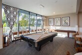 Located at 16700 Saybrook Ln in Huntington Beach, California, this single-story community clubhouse offers a thoughtfully designed gathering space with a coastal-inspired aesthetic. The main lounge area is anchored by a classic fireplace paired with a ceiling-mounted projector and retractable screen, making it ideal for both relaxed socializing and presentations. Rich parquet hardwood flooring runs throughout the lounge, complemented by comfortable upholstered seating and abundant natural light streaming through floor-to-ceiling glass walls that frame lush outdoor greenery and a pool area beyond. An adjacent sitting nook features elegant wainscoting, a decorative chandelier, and direct access to an outdoor patio through expansive sliding glass doors. The kitchen area showcases a generous granite-topped bar with bar stool seating, white cabinetry, a glass tile backsplash, and globe pendant lighting — well suited for catering or casual entertaining. Multiple dining zones accommodate various group sizes, with a mix of bistro-height tables and standard dining tables, all accented by coastal artwork and decorative mirrors. The open layout connects each area seamlessly, creating a versatile, welcoming environment that reflects the relaxed character of its Southern California waterfront community.