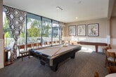 Located at 16700 Saybrook Ln in Huntington Beach, California, this single-story community clubhouse offers a thoughtfully designed gathering space with a coastal-inspired aesthetic. The main lounge area is anchored by a classic fireplace paired with a ceiling-mounted projector and retractable screen, making it ideal for both relaxed socializing and presentations. Rich parquet hardwood flooring runs throughout the lounge, complemented by comfortable upholstered seating and abundant natural light streaming through floor-to-ceiling glass walls that frame lush outdoor greenery and a pool area beyond. An adjacent sitting nook features elegant wainscoting, a decorative chandelier, and direct access to an outdoor patio through expansive sliding glass doors. The kitchen area showcases a generous granite-topped bar with bar stool seating, white cabinetry, a glass tile backsplash, and globe pendant lighting — well suited for catering or casual entertaining. Multiple dining zones accommodate various group sizes, with a mix of bistro-height tables and standard dining tables, all accented by coastal artwork and decorative mirrors. The open layout connects each area seamlessly, creating a versatile, welcoming environment that reflects the relaxed character of its Southern California waterfront community.