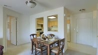 This single-floor residence offers a practical and comfortable layout centered around a combined dining and kitchen area. The dining space accommodates a traditional dark wood table surrounded by four cushioned chairs with floral upholstery, creating a welcoming setting for everyday meals. Soft carpeting underfoot and neutral white walls contribute to a clean, understated ambiance throughout.

A distinctive architectural feature separates yet connects the dining area to the kitchen — a partial wall with two open pass-through windows that allow natural sightlines into the cooking space. This design promotes an open feel while maintaining a defined kitchen zone. The kitchen is equipped with light wood cabinetry offering ample storage, a black gas range with an overhead range hood, a stainless steel refrigerator, and a sink positioned beneath the pass-through. Overhead flush-mount lighting in both the dining area and kitchen ensures consistent illumination throughout. A hallway visible to the right suggests additional living spaces beyond, contributing to the home's overall functional flow.