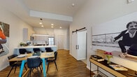 This thoughtfully designed single-floor residence showcases a seamless open-concept layout that connects the kitchen, dining, and living areas into one cohesive living space. The kitchen is appointed with dark espresso cabinetry, white quartz countertops, a subway tile backsplash, stainless steel appliances, and a peninsula with bar seating — all illuminated by recessed lighting and a sleek linear pendant fixture. Wood-look flooring flows throughout the main living areas, adding warmth and continuity. A sliding barn door serves as a stylish functional divider, lending an industrial-modern touch to the interior. The living area offers generous space and is bathed in natural light from large windows dressed with white blinds. The bedroom features plush carpeting, elevated ceilings, and bright windows that invite natural light. The full bathroom is finished with large-format tile, a soaking tub, a white quartz vanity with an undermount sink, and dark wood cabinetry. Throughout the space, high ceilings enhance the airy, open feel, while the neutral color palette and modern design elements create a clean, contemporary ambiance that is both inviting and functional.