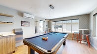 Located at 1946 Ambrosi Road in Kelowna, BC, this dedicated billiards room is a well-appointed game space designed for leisure and social interaction. At its center sits a full-size pool table with a blue felt surface, complemented by a sleek pendant light suspended directly overhead to ensure optimal table illumination. The room is finished with warm light wood flooring that contrasts tastefully with the patterned diamond-motif wallcovering, which lends the space a contemporary yet inviting character. A wall of large windows along one side floods the room with natural light and offers views of the surrounding Kelowna streetscape, while additional windows on an adjacent wall enhance the bright, open atmosphere. A wall-mounted cue rack keeps equipment neatly organized, and a dedicated area features high-top tables with modern stools, providing a comfortable spot for spectators. Climate control is handled by a wall-mounted split unit, ensuring year-round comfort. A wall-mounted flat-screen television and decorative artwork add personality to the space. Recessed ceiling lighting supplements the pendant fixture for well-balanced illumination throughout. The room is accessed through a set of glass-panel doors, maintaining a modern aesthetic while connecting seamlessly to the broader facility.