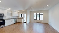 This single-floor space showcases a clean, contemporary design centered around an open-concept living and kitchen area. Light-toned hardwood flooring flows seamlessly throughout, lending warmth and continuity to the layout. The kitchen is outfitted with white shaker-style cabinetry, dark countertops, and stainless steel appliances, including a dishwasher. Recessed lighting is installed throughout the ceiling, providing consistent, even illumination that complements the abundant natural light streaming in through multiple black-framed windows. These windows, positioned along two walls, frame views of the surrounding exterior and contribute to an airy, well-lit ambiance. The open living area is generously proportioned, offering flexible space for a variety of furniture arrangements. Crisp white walls and clean architectural lines reinforce the modern aesthetic. A glass-paneled door near the kitchen provides additional light and access to an outdoor area. Overall, the space presents a fresh, move-in-ready environment with a minimalist sensibility and a bright, welcoming atmosphere suited to a variety of uses.