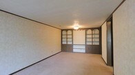 This single-floor space presents a straightforward, functional layout with a notably classic design aesthetic. The main living area is carpeted in a neutral beige tone that extends throughout, creating a cohesive and understated foundation. The walls are finished with a subtle textured wallcovering trimmed with dark wood crown molding and baseboards, adding a traditional character to the interior.

The room's standout feature is a built-in wall unit spanning the full width of one wall. This dark wood shelving system includes multiple open compartments framed by decorative arched detailing at the top, providing generous display and storage space. Cabinet doors at the lower level offer concealed storage, while a central recessed section appears designed to accommodate a television or media display. Overhead, a flush-mount ceiling light fixture provides general illumination beneath a textured ceiling. The overall ambiance is calm and neutral, with the dark wood built-ins serving as a strong visual anchor against the lighter surrounding surfaces. The space is well-suited for use as a living or family room, offering both storage functionality and a classic, timeless character.