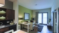 This single-story living space is designed with a contemporary aesthetic that balances bold color choices with clean, modern lines. The focal point of the main living area is a striking green accent wall, which adds a lively pop of color and provides a dynamic backdrop for the framed artwork displayed upon it. The remaining walls are finished in a soft neutral tone, creating a balanced contrast that keeps the space feeling open and inviting.

The living area is furnished with a light-toned sofa accented by decorative pillows in earthy tones, complemented by a geometric-patterned area rug that anchors the seating arrangement. A streamlined white console table serves as both a functional and decorative element, styled with candles and a table lamp that contributes to the warm ambiance. An additional lamp provides layered lighting throughout the space.

A sliding glass door with floor-length drapes allows abundant natural light to flood the interior while offering access to the outdoors. The tiled flooring throughout is practical and cohesive, tying the space together. Dark cabinetry visible toward the perimeter suggests additional storage or a nearby functional area, adding utility to the overall layout.