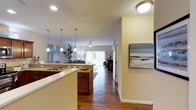 This single-floor residence showcases a thoughtfully designed open-concept layout that seamlessly connects the kitchen, dining, and living areas. The kitchen is appointed with rich dark wood cabinetry, granite countertops, a double basin sink, and an island with elegant pendant lighting overhead — creating both a functional and visually appealing workspace. Warm hardwood flooring flows continuously throughout, tying the spaces together cohesively.

The living room is generously proportioned and filled with natural light, courtesy of large sliding glass doors that offer views of the outdoors. A ceiling fan anchors the room above, while a textured ceiling adds architectural character. The space comfortably accommodates a full seating arrangement, making it ideal for both relaxing and entertaining.

A hallway lined with soft neutral walls connects the main living areas, with recessed and flush-mount lighting ensuring the home remains bright throughout. Tasteful coastal artwork adorns the walls, enhancing the welcoming ambiance. Overall, this home offers a clean, comfortable, and well-lit environment with a warm, inviting aesthetic supported by quality finishes and an efficient, flowing floor plan.