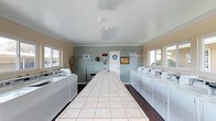 This single-floor communal laundry facility is designed for efficiency and ease of use. The room is laid out with rows of full-sized washing machines and dryers lining both side walls, providing ample capacity for residents. At the center of the space sits a long, tiled folding table, offering a generous surface for sorting and folding laundry. The room features dark hardwood-style flooring that contrasts cleanly with the light-colored walls and white appliances. Crown molding adds a subtle finishing touch to the ceiling line, lending the space a polished appearance. Multiple windows along the walls invite natural light, keeping the room bright and open. A utility sink is conveniently positioned near the back wall, and a doorway provides additional access. Informational signage and a bulletin board are mounted on the walls, keeping users informed of facility rules and notices. A smoke detector is visible on the ceiling, reflecting attention to safety. Overall, the space is clean, functional, and thoughtfully organized to serve the practical needs of its users.