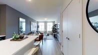 This single-floor space showcases a thoughtfully designed open-concept layout that connects the dining and living areas in one cohesive flow. Light grey hardwood-style flooring runs throughout, complementing the cool-toned slate-grey accent walls and crisp white ceilings that lend the space a clean, contemporary aesthetic.

A white kitchen peninsula anchors the dining area, paired with modern bar-style seating in brown leather and white finishes. The peninsula provides practical counter space while naturally defining the boundary between the kitchen and the rest of the living area.

The living area is well-appointed with a comfortable seating arrangement centered around a light area rug. A sleek black open shelving unit serves as a media and display wall, housing a flat-screen television alongside decorative accents. Natural light filters generously into the space through multiple windows dressed with sheer white curtains, brightening the interior and creating an airy ambiance.

A large white door on the right side of the space leads to an additional room, suggesting a private area beyond the main open-concept zone. Overall, the space presents a modern, functional design with a neutral palette and clean architectural lines.