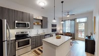 This well-appointed single-floor residence offers a thoughtfully designed open-concept layout that seamlessly connects the kitchen and living areas. The kitchen is a standout feature, showcasing dark wood-toned cabinetry, stainless steel appliances including a range, microwave, dishwasher, and refrigerator, and a granite-topped island that serves as both a prep space and casual gathering point. Stylish pendant lighting above the island adds a modern touch, while light wood-style flooring flows throughout the main living areas, enhancing the bright, airy atmosphere. The living area enjoys abundant natural light through large windows dressed with elegant draping curtains and blinds, and a ceiling fan provides added comfort. A private exterior access door is also visible from the living space. The bedrooms are carpeted for warmth and comfort, each benefiting from natural light through well-appointed windows. The primary bedroom features its own ceiling fan and direct access to an outdoor space. A walk-in closet with built-in wire shelving provides generous storage. Clean white walls and understated finishes throughout create a neutral, welcoming canvas that suits a variety of interior styles.